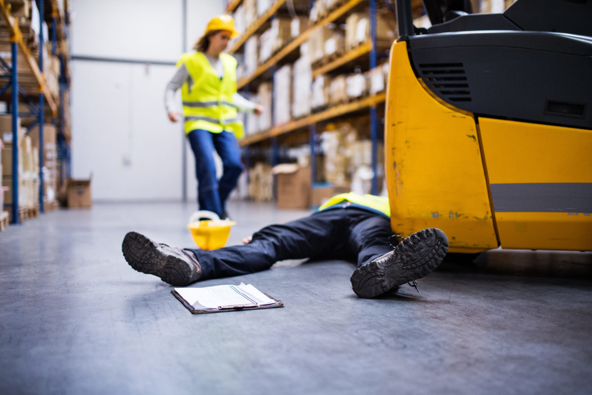 An accident in a warehouse. Woman running towards her colleague lying on the floor next to a forklift.