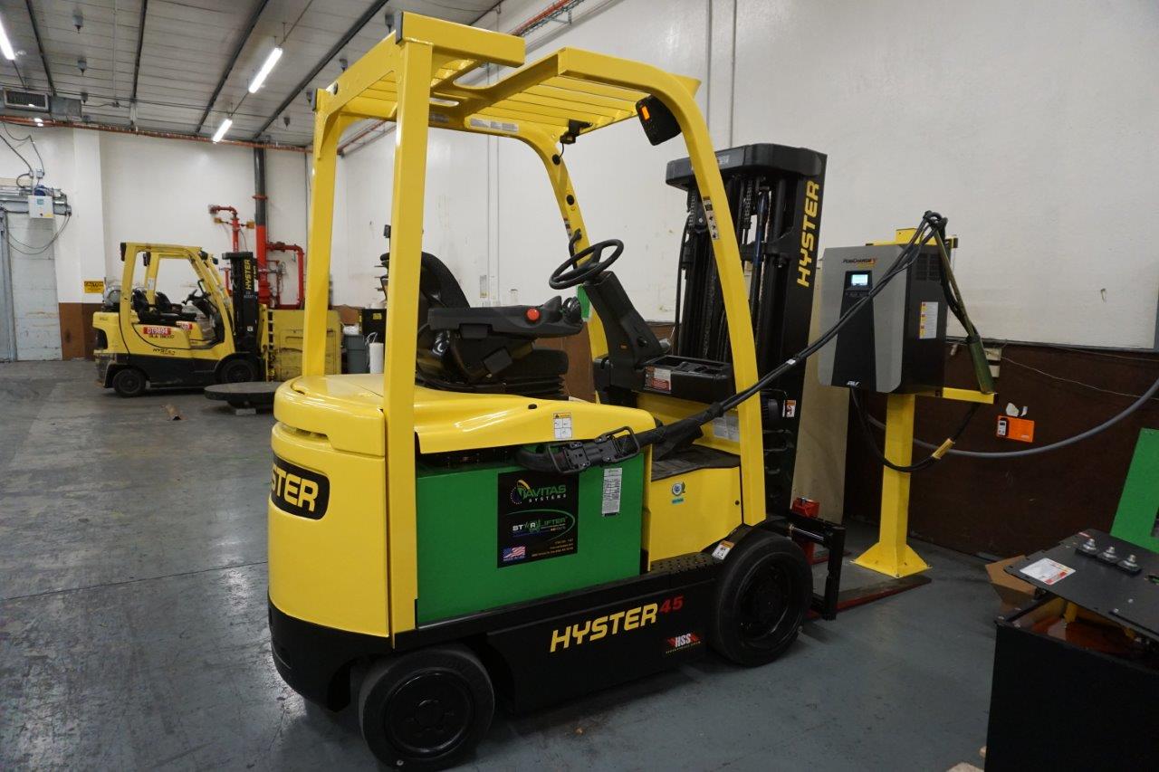 Navitas-Systems-Starlifter-Forklift-connected-to-charger-foreground-propane-forklifts-background